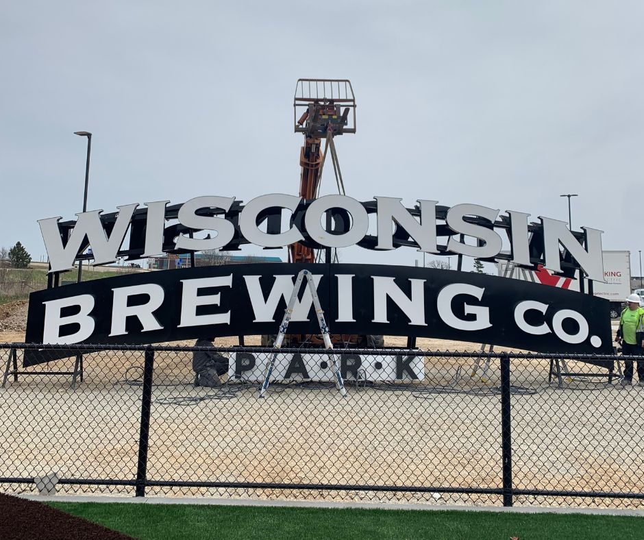 Image includes a large exterior sign with the words "Wisconsin Brewing Co." in large letters. Instatllation bucket truck is behind the sign, and is lowering the sign into place.