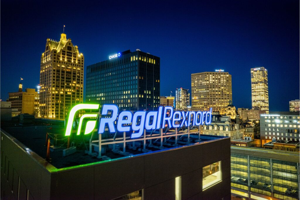 A night time image of the Regal Rexnord channel letter sign. The sign includes a green illuminated logo with the words "Regal Rexnord" in blue. The cityscape of Milwaukee is in the background.