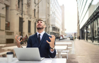 A person sitting at an outdoor desk, wringing his hands in frustration while looking upward.